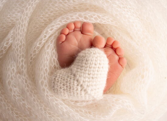 Nahaufnahme von Babyfüßen, die in einem weißen, weichen Tuch eingewickelt sind, mit einem gestrickten weißen Herz zwischen den Füßen