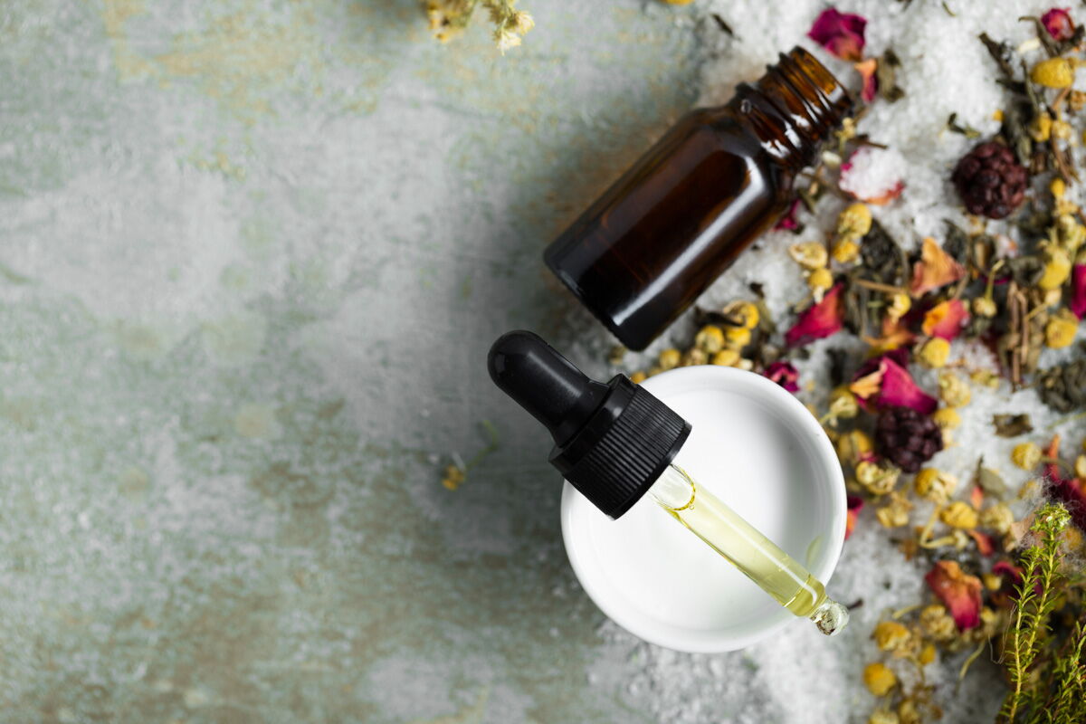 Braune Glasflasche mit Pipette und gelblichem Serum, umgeben von getrockneten Blüten und Kräutern auf hellem Untergrund